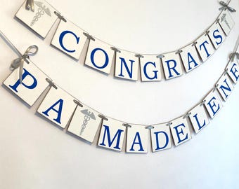 Puede incluir: Pancarta blanca con letras azules que deletrean "CONGRATS MADELEINE" y un símbolo médico plateado en cada extremo.