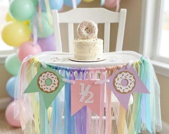 May include: A small white frosted cake topped with a donut sits on a high chair tray. A pastel banner hangs from the tray, featuring two donut illustrations and a pink center flag with the white text 1/2, accented by hanging tulle ribbons.