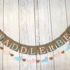 May include: A gender reveal banner with the text "WADDLE it BE?" in brown, pink, and blue letters. The banner is decorated with pink and blue duck silhouettes and hearts.