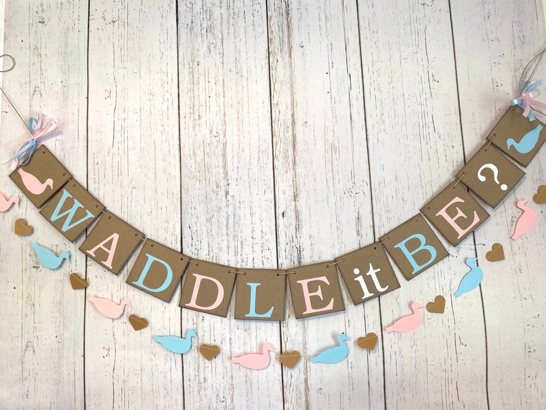 May include: A gender reveal banner with the text "WADDLE it BE?" in brown, pink, and blue letters. The banner is decorated with pink and blue duck silhouettes and hearts.