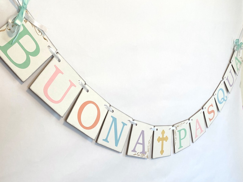 May include: A white banner with the words "BUONA PASQUA" in pastel colors. The letters are on individual squares with a gold cross in between the words. The banner is strung with white ribbon.