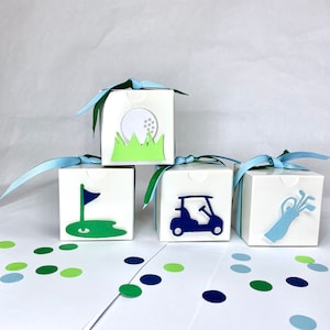May include: Four white favor boxes with blue ribbon tied around them. Each box has a different golf-themed design: a golf ball, a golf flag, a golf cart, and a golf bag. The boxes are on a white surface with green and blue confetti.