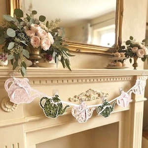 May include: A decorative garland featuring five paper teapot cutouts strung on a white ribbon. The teapots alternate between a light pink floral pattern and a dark green botanical print, each accented with a small white ribbon bow on the handle.