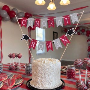 May include: A white frosted cake topped with a two-tiered banner on wooden skewers. The top banner reads CONGRATS in white letters on red and gray pennants. The bottom banner features black graduation caps and spells ROWAN in white letters.