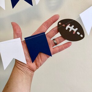 May include: A white and navy blue paper banner with brown football cutouts. The banner is strung together with white thread.