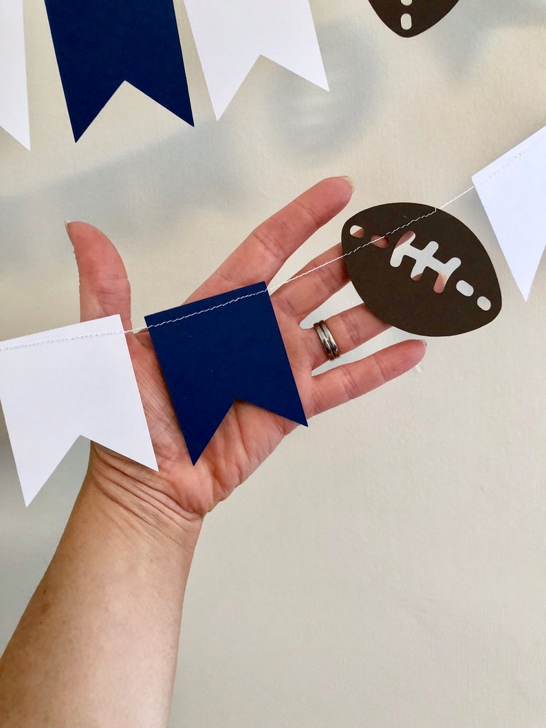 May include: A white and navy blue paper banner with brown football cutouts. The banner is strung together with white thread.