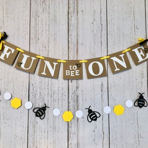 Puede incluir: Una pancarta marr&oacute;n y blanca con el texto "Fun to Bee One" y decoraciones de abejas amarillas y negras. Una cadena de c&iacute;rculos blancos, hex&aacute;gonos amarillos y recortes de abejas negras cuelga debajo de la pancarta.