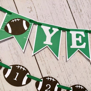 May include: Green and white banner with the words "First Year" and football graphics. The banner is strung with green ribbon.