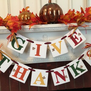 K&ouml;nnte beinhalten: Ein Thanksgiving-Banner mit den Worten "Give Thanks" in roten, gr&uuml;nen und gelben Buchstaben auf wei&szlig;en Quadraten. Das Banner ist mit Herbstbl&auml;ttern und einem kleinen K&uuml;rbis verziert.