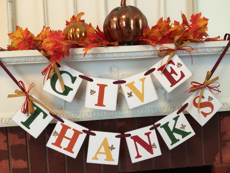 K&ouml;nnte beinhalten: Ein Thanksgiving-Banner mit den Worten "Give Thanks" in roten, gr&uuml;nen und gelben Buchstaben auf wei&szlig;en Quadraten. Das Banner ist mit Herbstbl&auml;ttern und einem kleinen K&uuml;rbis verziert.