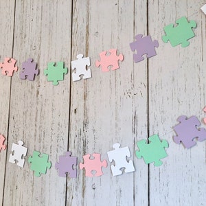 May include: A garland of pastel colored paper puzzle pieces strung together with white thread. The garland is made of light pink, light green, light purple, and white puzzle pieces.