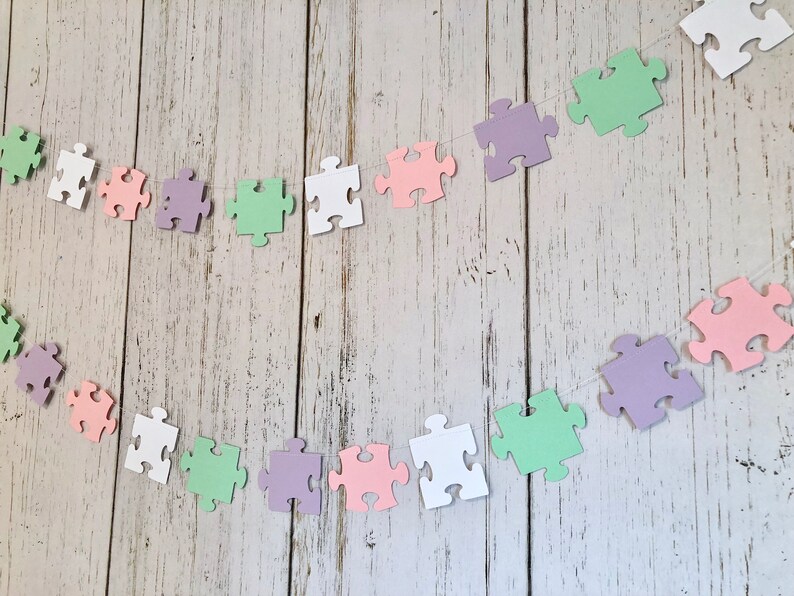 May include: A garland of pastel colored paper puzzle pieces strung together with white thread. The garland is made of light pink, light green, light purple, and white puzzle pieces.