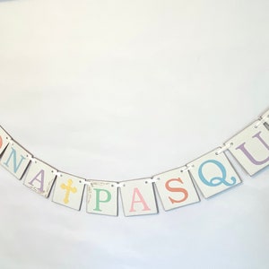 May include: A white banner with the words "BUONA PASQUA" in pastel colors. Each letter is on a separate square with a white background and a gold border. The banner is strung with white ribbon and has a cross on the "A".
