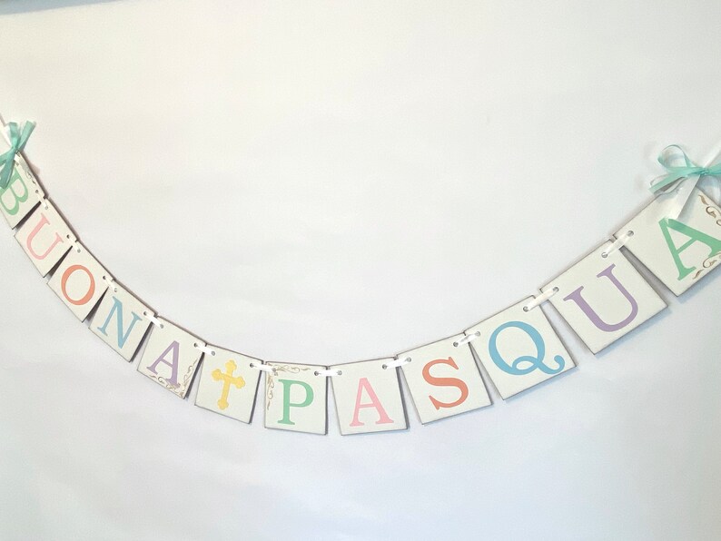 May include: A white banner with the words "BUONA PASQUA" in pastel colors. Each letter is on a separate square with a white background and a gold border. The banner is strung with white ribbon and has a cross on the "A".