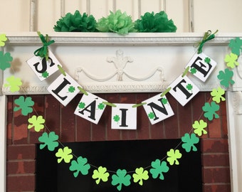 May include: A green shamrock garland with a white banner that reads "Sláinte" in black letters. The garland is hanging above a brick fireplace.