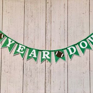May include: Green and white banner with the text "1st Year Down" and football graphics. The banner is strung on a white wooden background.