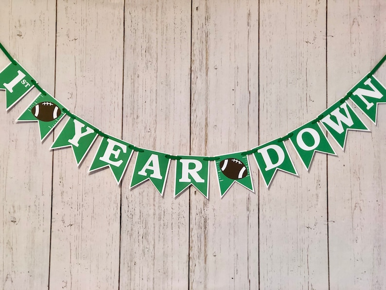 May include: Green and white banner with the text "1st Year Down" and football graphics. The banner is strung on a white wooden background.