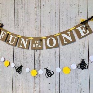 Puede incluir: Una pancarta de papel marr&oacute;n con el texto "Fun to Bee One" en letras blancas. La pancarta est&aacute; decorada con abejas amarillas y negras y est&aacute; atada con una cinta amarilla. Una cadena separada de abejas negras, c&iacute;rculos blancos y hex&aacute;gonos amarillos cuelga debajo de la pancarta.