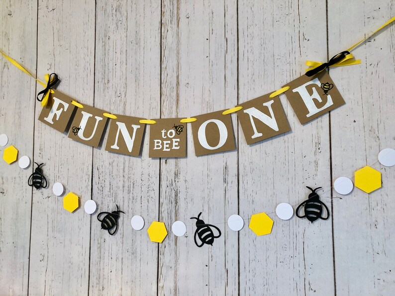 Puede incluir: Una pancarta de papel marr&oacute;n con el texto "Fun to Bee One" en letras blancas. La pancarta est&aacute; decorada con abejas amarillas y negras y est&aacute; atada con una cinta amarilla. Una cadena separada de abejas negras, c&iacute;rculos blancos y hex&aacute;gonos amarillos cuelga debajo de la pancarta.