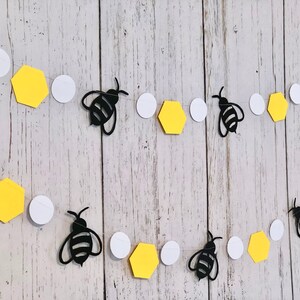 Puede incluir: Una guirnalda de abejas y panales negros y blancos con panales amarillos y c&iacute;rculos blancos. La guirnalda est&aacute; enhebrada en una cuerda transparente.