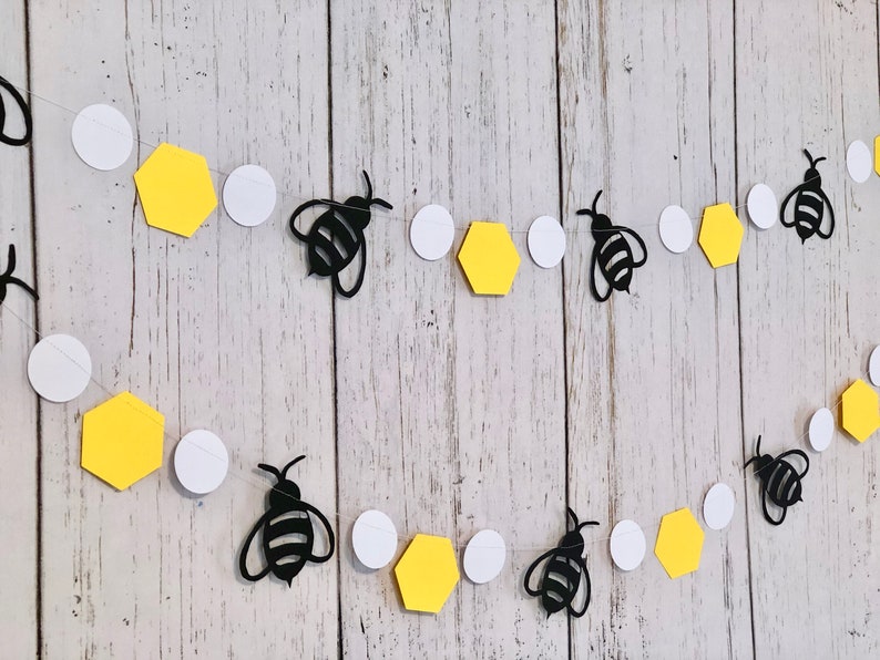 Puede incluir: Una guirnalda de abejas y panales negros y blancos con panales amarillos y c&iacute;rculos blancos. La guirnalda est&aacute; enhebrada en una cuerda transparente.