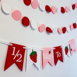 May include: A festive banner with red, white, and pink circular garland. Below, pennant flags spell out "1/2 Way To One" with strawberry illustrations. Perfect for a birthday celebration.