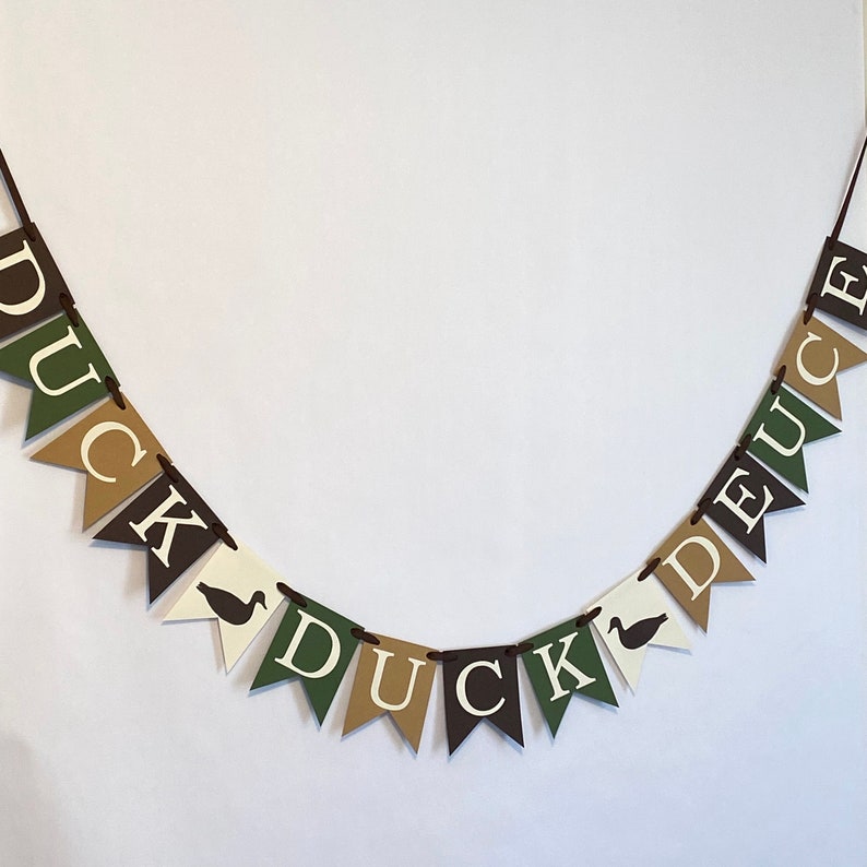 May include: A brown, green, and white banner with the word "DUCK" on one side and "DUCK" on the other side. The banner is made of felt and has a string for hanging.