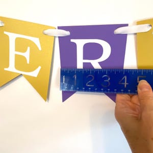 May include: A purple and gold banner with the word "FRI" in white letters. The banner is being measured with a ruler, showing that each letter is approximately 5 inches wide.