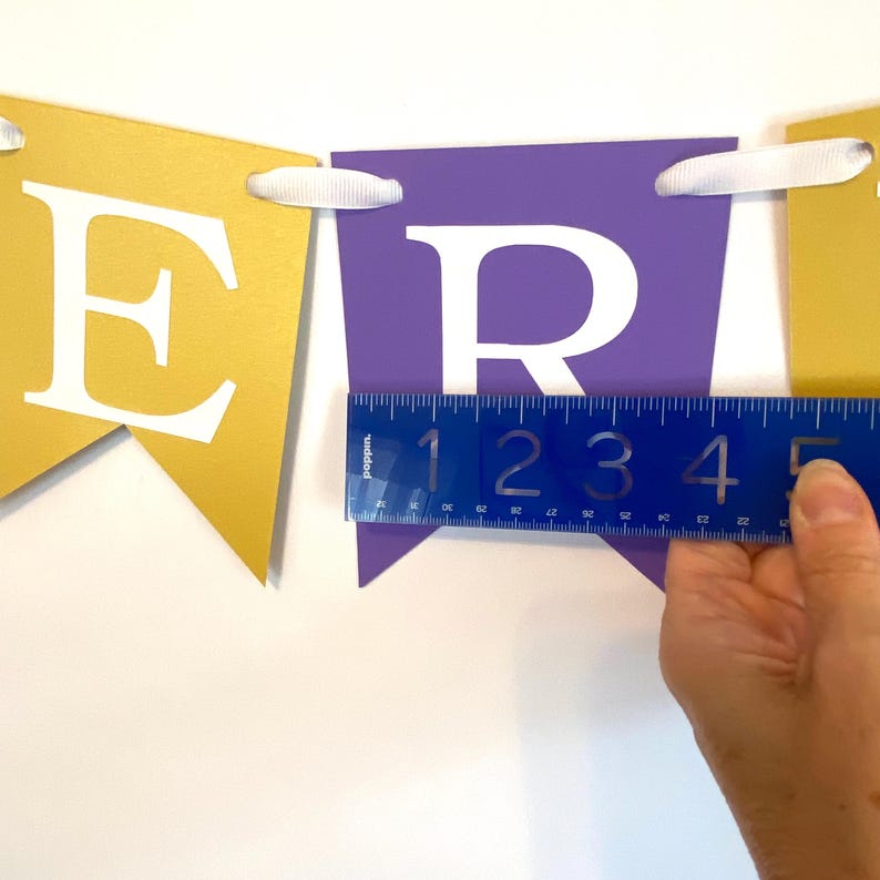 May include: A purple and gold banner with the word "FRI" in white letters. The banner is being measured with a ruler, showing that each letter is approximately 5 inches wide.