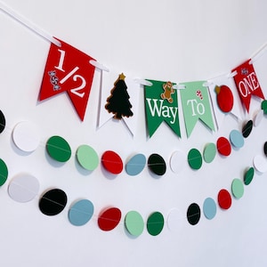 May include: A festive Christmas banner with red and green pennants, featuring the text "1/2 Way To One". The banner includes a Christmas tree, gingerbread man, and candy cane decorations. Two strands of colorful circles complete the holiday decor.