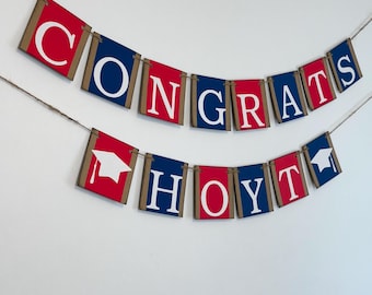 May include: A two-tiered graduation banner featuring rectangular panels in red and navy blue with white lettering spelling out CONGRATS HOYT. The panels are strung together on twine, with white graduation cap icons on the first and last panels.