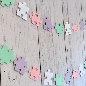 May include: A garland of pastel colored paper puzzle pieces strung together with white thread. The garland is made up of white, light pink, light green, and light purple puzzle pieces.