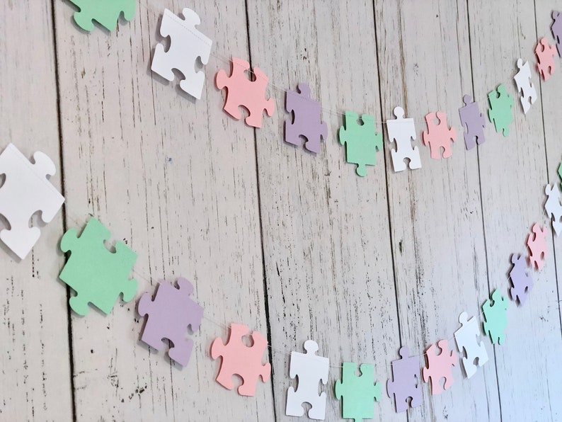 May include: A garland of pastel colored paper puzzle pieces strung together with white thread. The garland is made up of white, light pink, light green, and light purple puzzle pieces.