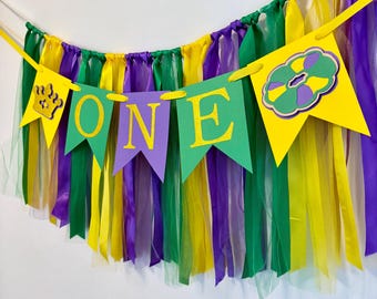 May include: A Mardi Gras themed banner with the word "ONE" in gold glitter letters on green and purple flags. The banner is decorated with yellow, green, and purple streamers and a purple and green donut.