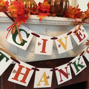 K&ouml;nnte beinhalten: Ein festliches Thanksgiving-Banner mit den Worten "Give Thanks" in roten, gr&uuml;nen, orangefarbenen und gelben Buchstaben. Das Banner ist mit kleinen goldenen Blattakzenten verziert und mit einem burgunderfarbenen Band befestigt.