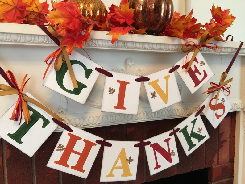 K&ouml;nnte beinhalten: Ein festliches Thanksgiving-Banner mit den Worten "Give Thanks" in roten, gr&uuml;nen, orangefarbenen und gelben Buchstaben. Das Banner ist mit kleinen goldenen Blattakzenten verziert und mit einem burgunderfarbenen Band befestigt.