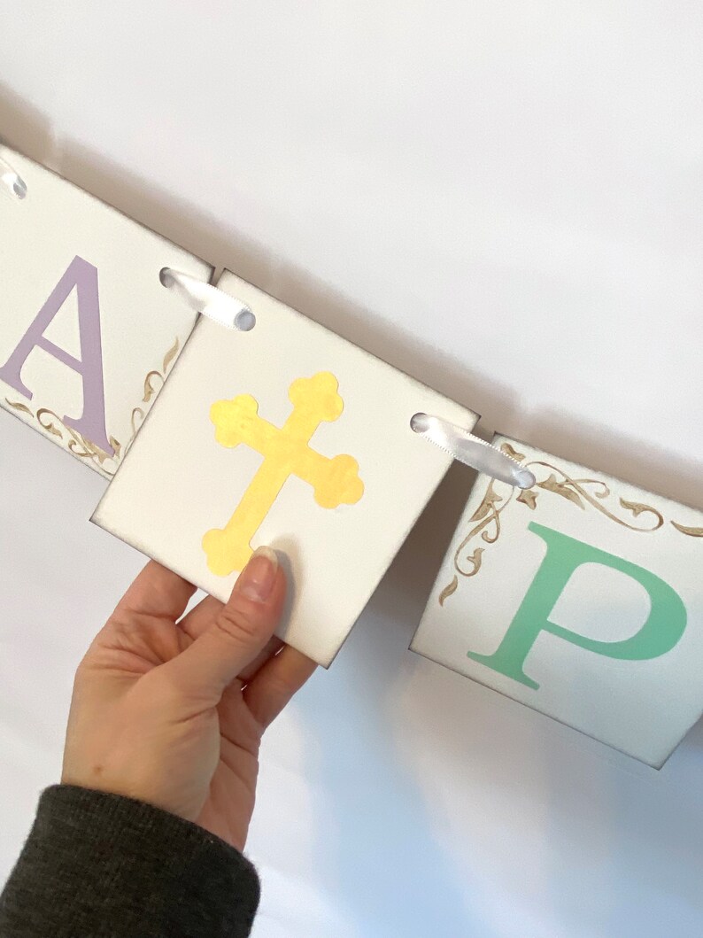 May include: A white banner with the word "Happy" in purple, a gold cross, and the letter "P" in green. The banner is strung together with white ribbon.