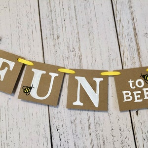 Puede incluir: Una pancarta de papel marr&oacute;n con las palabras "FUN to BEE" en letras blancas. La pancarta est&aacute; decorada con una cuerda amarilla y peque&ntilde;as ilustraciones de abejas.