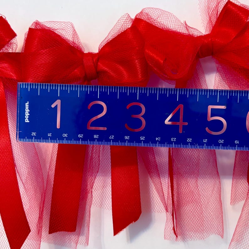 May include: Red tulle and satin ribbon bows with frayed edges, measured with a blue ruler marked with numbers 1 through 5. The ruler is 5 inches long.