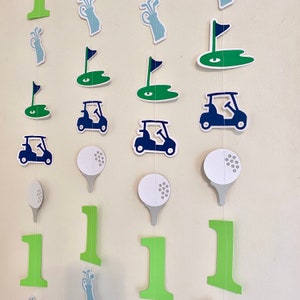 May include: A green, blue, and white golf-themed garland with golf carts, golf balls, golf clubs, and the number one. The garland is made of paper and is perfect for a golf-themed party.