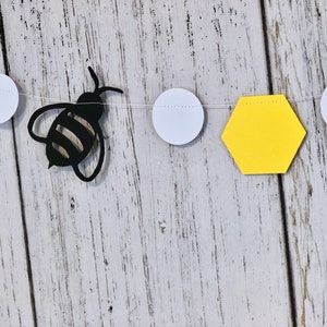 Puede incluir: Un recorte de abeja de papel negro con rayas amarillas y negras cuelga de una cuerda blanca con recortes de hex&aacute;gonos amarillos y c&iacute;rculos blancos. La cuerda cuelga sobre un fondo de madera blanca.