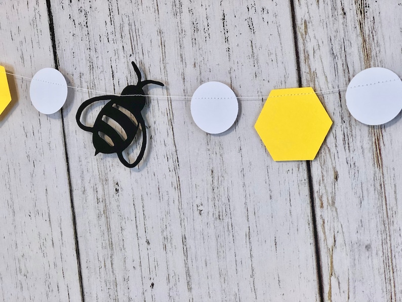 Puede incluir: Un recorte de abeja de papel negro con rayas amarillas y negras cuelga de una cuerda blanca con recortes de hex&aacute;gonos amarillos y c&iacute;rculos blancos. La cuerda cuelga sobre un fondo de madera blanca.