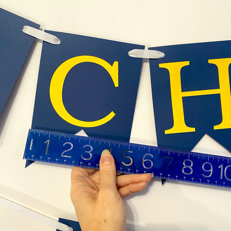 May include: A blue and yellow banner with the word "CHEERS" spelled out. The banner is made of felt and has white ribbon ties. A ruler is used to measure the banner, which is approximately 10 inches long.