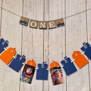 May include: A festive banner with the word "ONE" and truck illustrations, strung with orange and navy blue monthly photo holders numbered 1-12. Each photo holder has a pumpkin shape at the top. The banner is designed for a first birthday celebration.