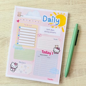May include: A rectangular daily planner notepad with a light pink background, featuring a yellow sun, clouds, and Hello Kitty illustrations. It includes sections for a to-do list, goals, appointments, and notes, with the text Daily Planner and Soft days, strong dreams printed on the page. A light green pen is placed next to the notepad.