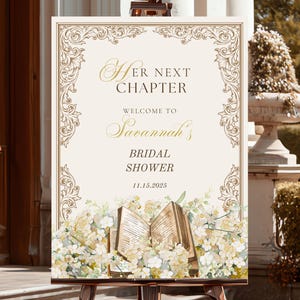 May include: A decorative sign on a wooden easel, featuring the text "Her Next Chapter" and "Welcome to Savannah's Bridal Shower 11.15.2025". The sign has a floral and ornate border design with an open book illustration.