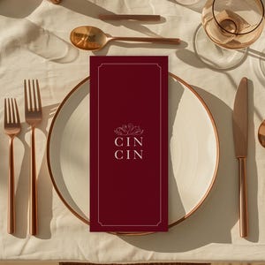 May include: Elegant table setting with copper-toned cutlery, candle holders, and glassware. A burgundy menu card with the text "CIN CIN" sits on a white plate. The scene is set on a cream-colored tablecloth, creating a warm, inviting atmosphere.