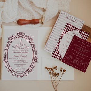 May include: Wedding invitation suite featuring a large invitation with an oval frame and swan illustration, plus smaller cards with burgundy and white designs. A wooden stamp and white fabric are also in the image.