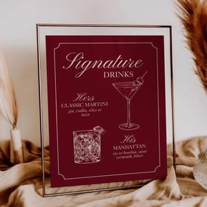 May include: A framed burgundy sign with the words "Signature Drinks" in elegant script. The sign features illustrations of a martini and an old-fashioned cocktail, with text indicating drink ingredients. Decorative dried grasses in a vase are in the background.
