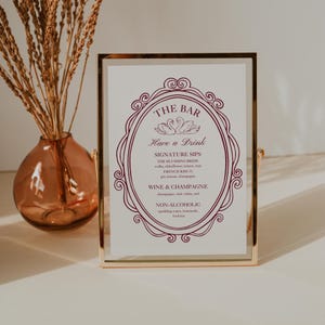 May include: A framed sign with the text "THE BAR" and "Have a Drink" in a decorative oval frame. The sign lists signature drinks, wine, champagne, and non-alcoholic options. The frame is gold-colored, and the background is white.